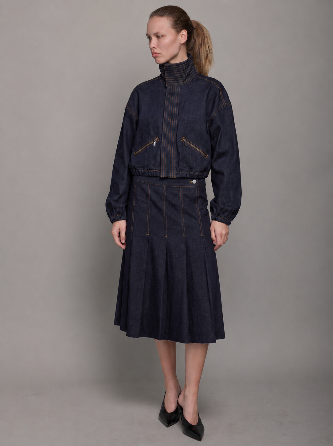 woman wearing a dark denim pleated midi skirt with a dark denim funnel neck jacket.