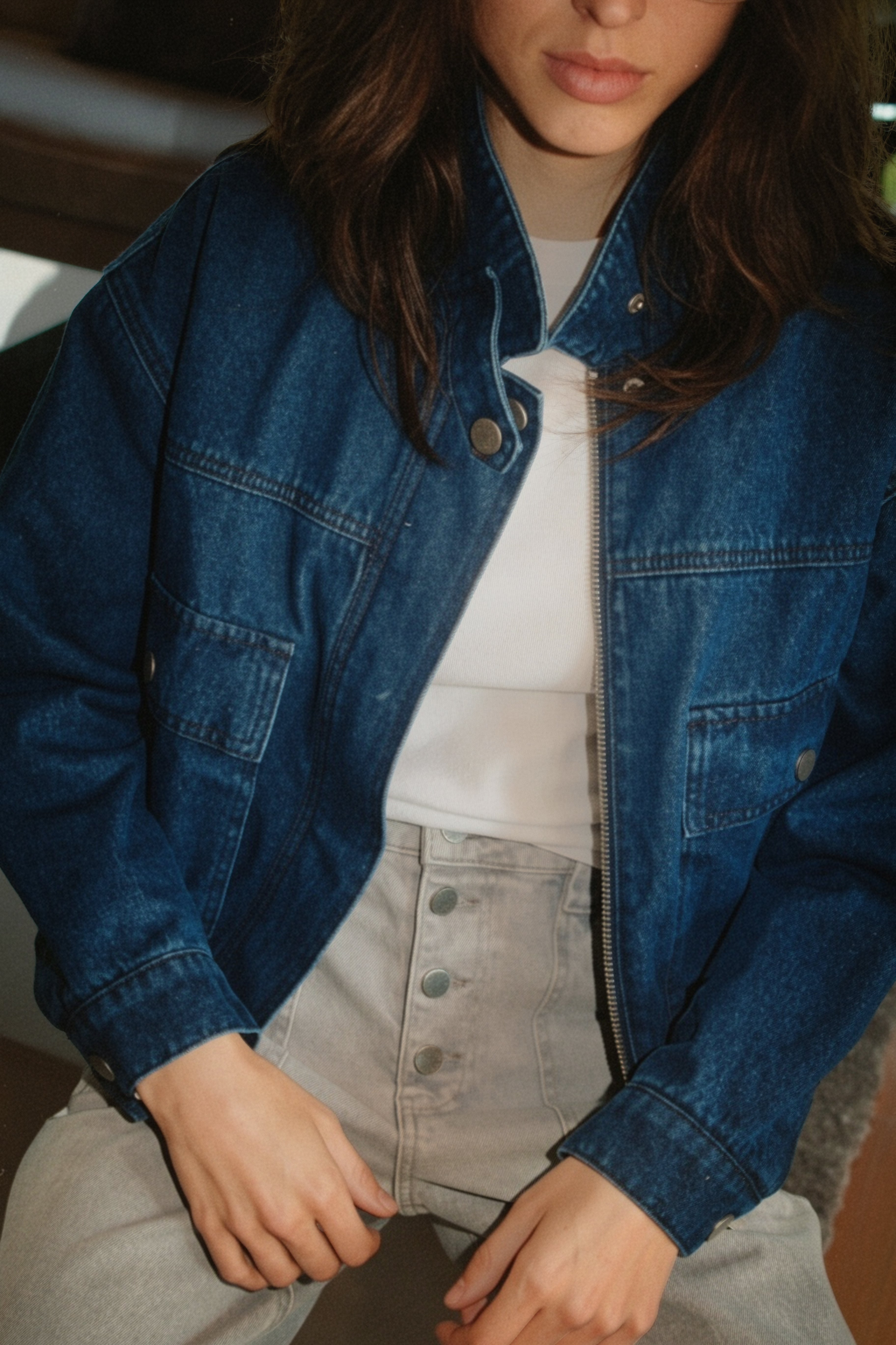 A close-up of a person wearing a blue denim jacket over a white top and light gray high-waisted button-front jeans.