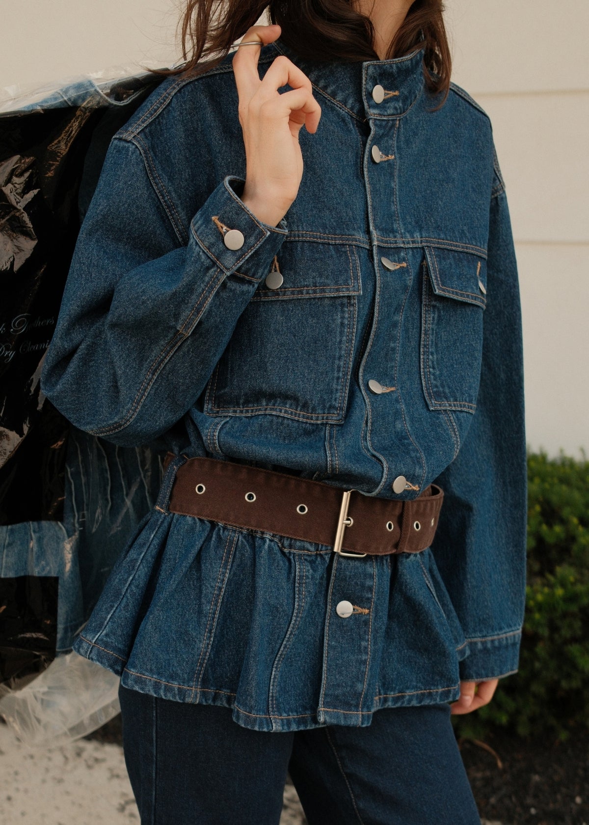 A person wearing an oversized blue denim jacket with front flap pockets and a brown belt at the waist, paired with dark jeans, photographed outdoors against a light wall and greenery.