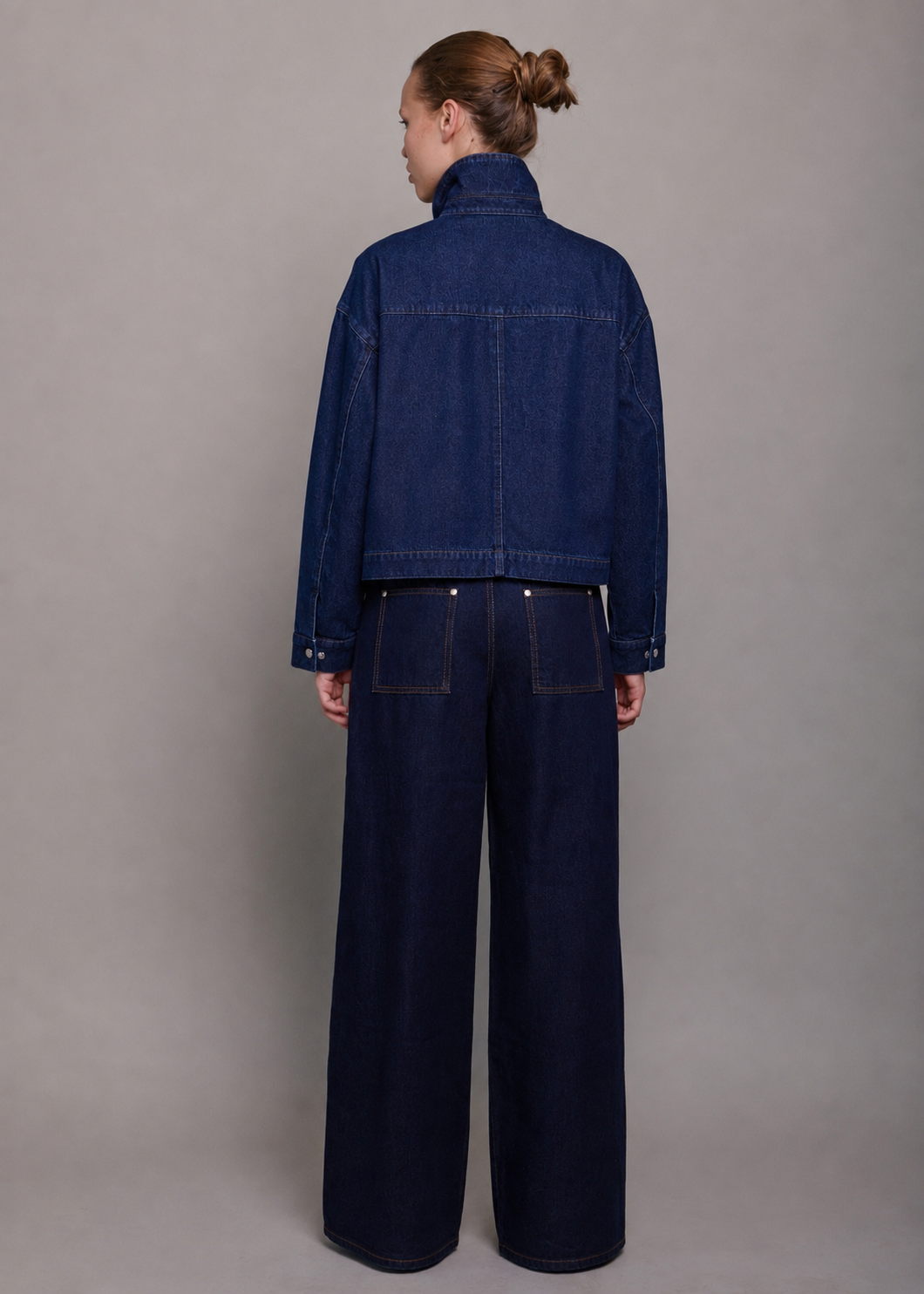 A model viewed from the back wearing a dark blue cropped denim jacket and matching wide-leg denim pants against a neutral gray studio background.
