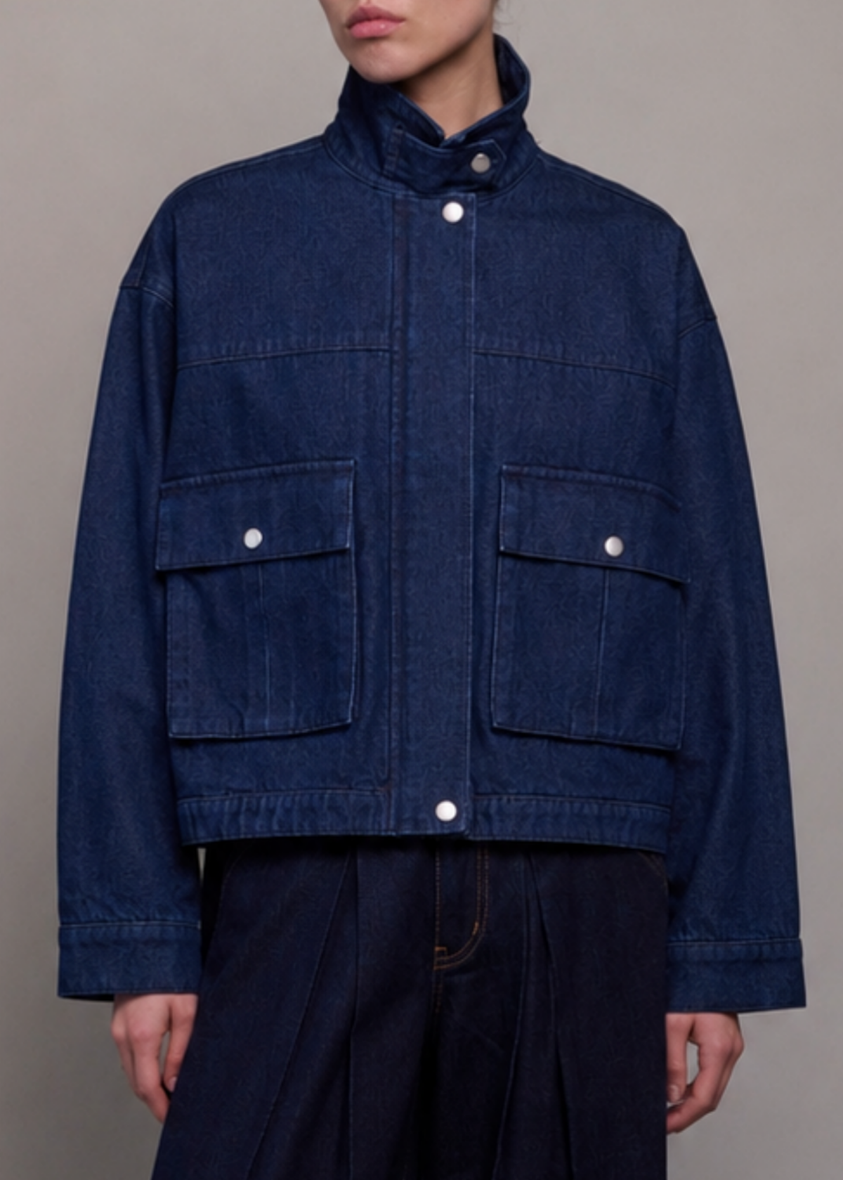 A person wearing a dark indigo denim jacket with a high stand collar, snap-button front, and two large flap chest pockets, paired with dark wide-leg pants against a plain light gray studio background.