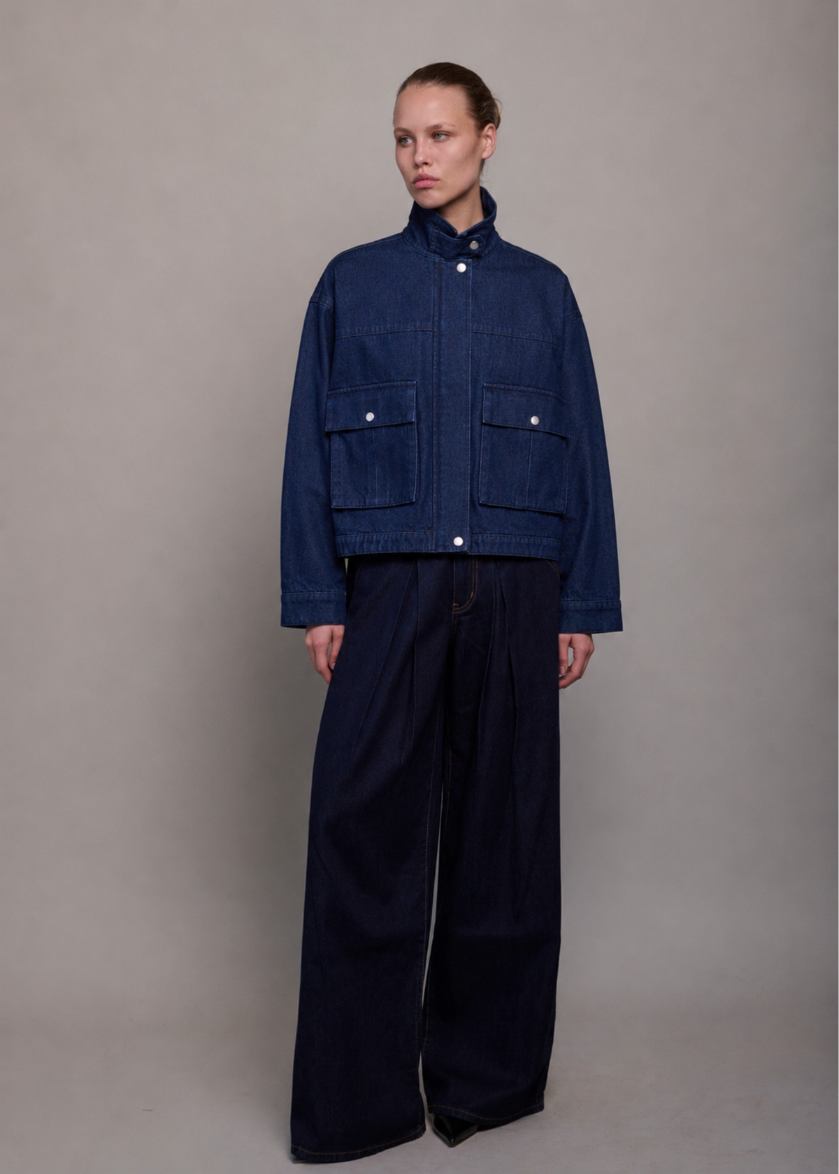 A model stands against a neutral gray studio background wearing a dark indigo denim jacket with a high collar and front flap pockets, paired with matching dark wide-leg trousers.