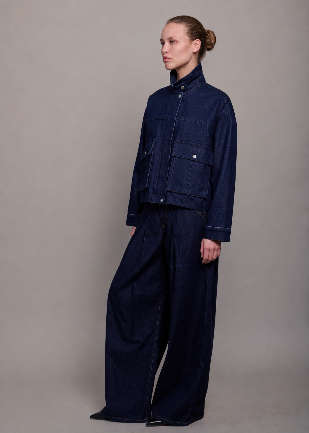 A model wearing a dark indigo denim jacket and matching wide-leg pants, shown in profile against a neutral gray studio background.