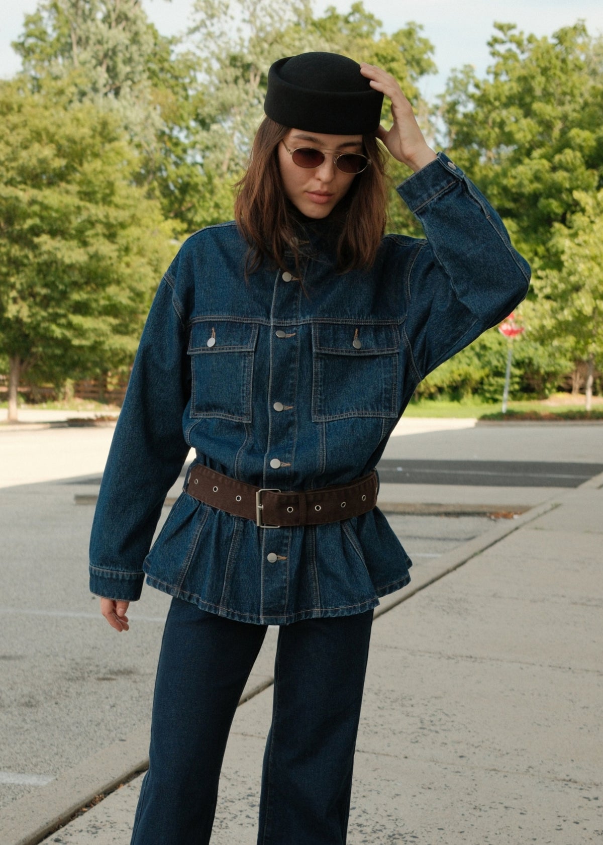 A woman wearing a dark blue denim jacket with a peplum waist, a brown studded belt, dark blue jeans, a black hat, and sunglasses, standing outdoors on a paved area with trees in the background.