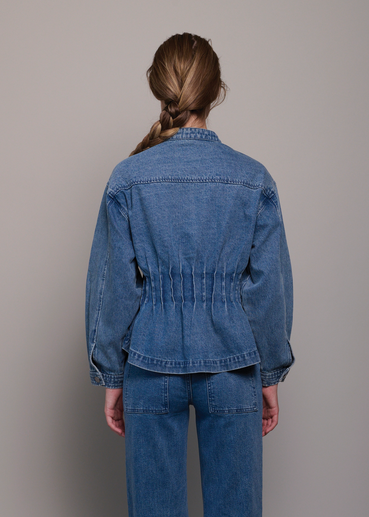 A model shown from the back wearing a blue denim jacket with gathered detailing at the waist and matching blue denim jeans, set against a plain gray studio background.