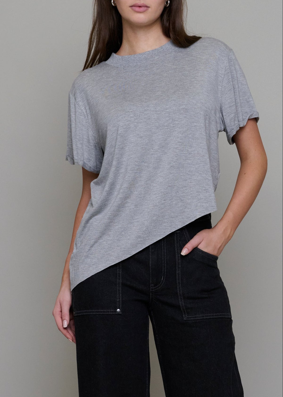 A woman wearing a light gray heathered short-sleeve t-shirt with a diagonal hem and black wide-leg jeans, standing against a solid gray studio background.