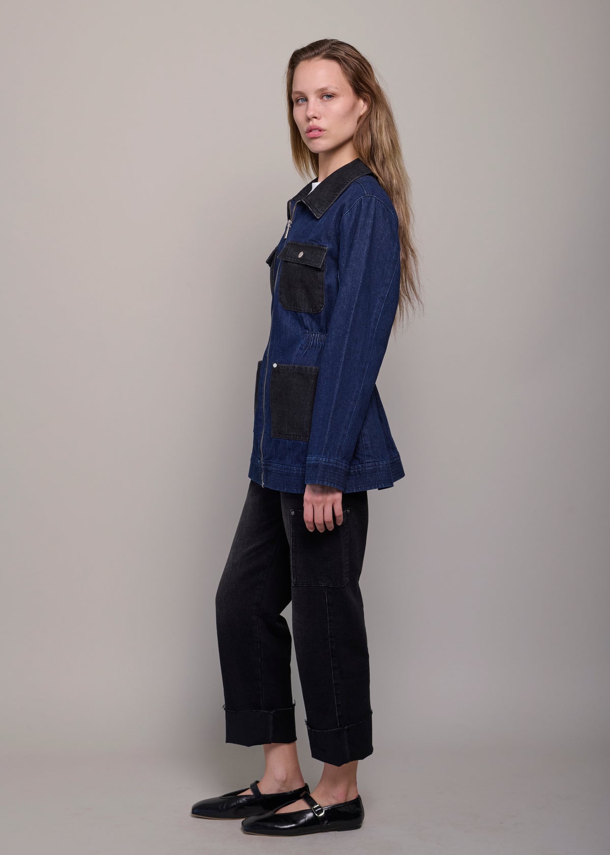 A woman modeling a dark indigo denim jacket with black contrast panels on the collar, chest pocket, and lower front pocket, paired with black cropped pants and black Mary Jane shoes, against a neutral gray studio background.