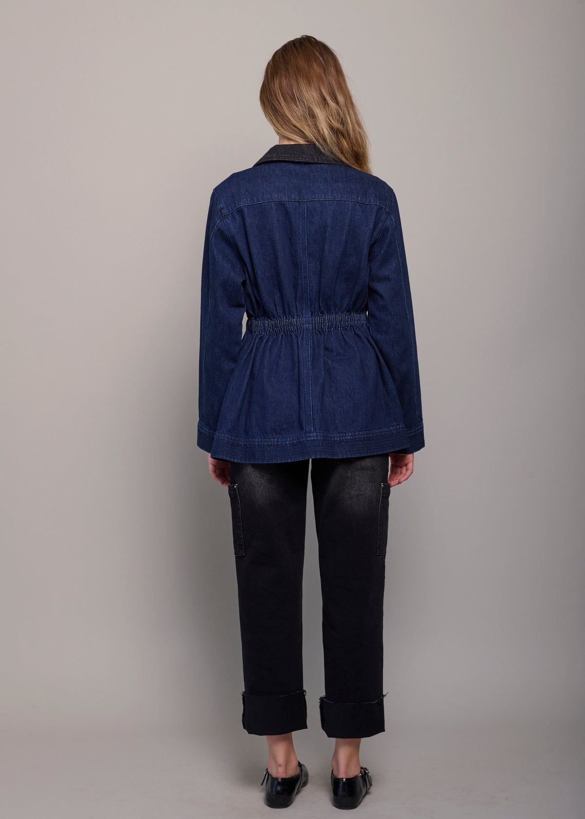 A woman viewed from the back wearing a dark blue denim belted jacket, dark cuffed jeans, and black shoes against a plain light gray studio background.
