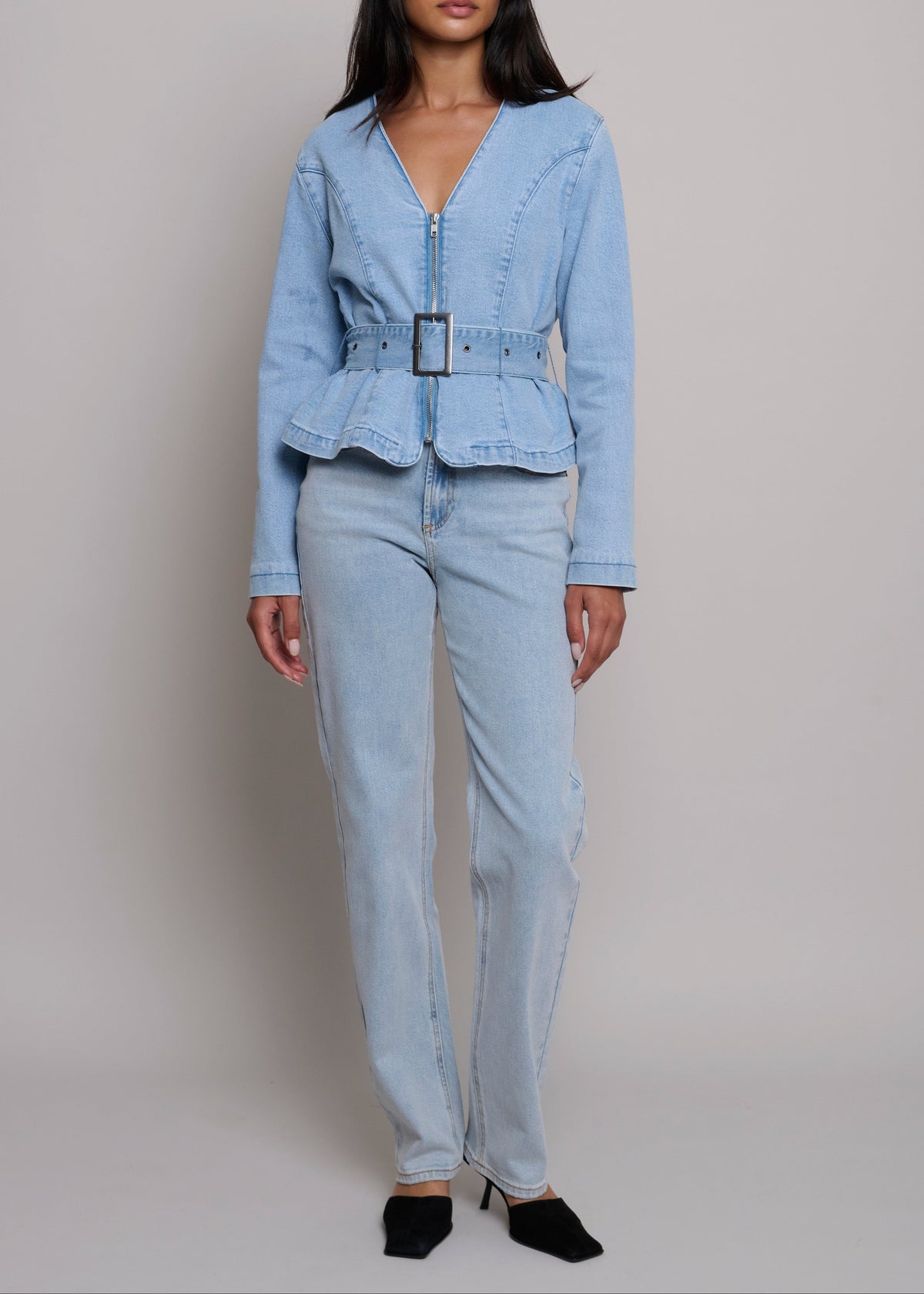 A model wearing a light blue denim two-piece set consisting of a long-sleeve belted jacket and matching straight-leg jeans, paired with black pointed-toe heels against a neutral studio background.