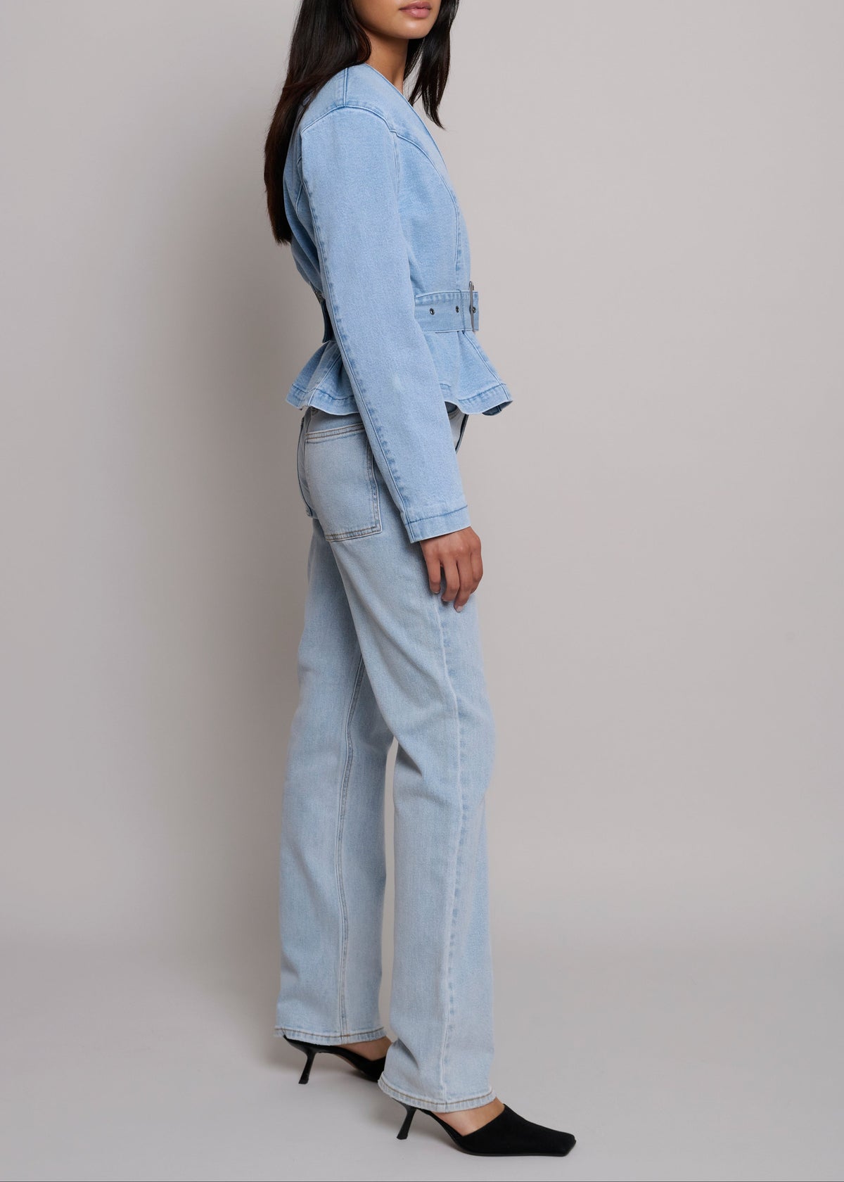A model shown from the side wearing a light blue denim jacket and matching wide-leg jeans, paired with black pointed-toe heels, against a neutral light gray studio background.