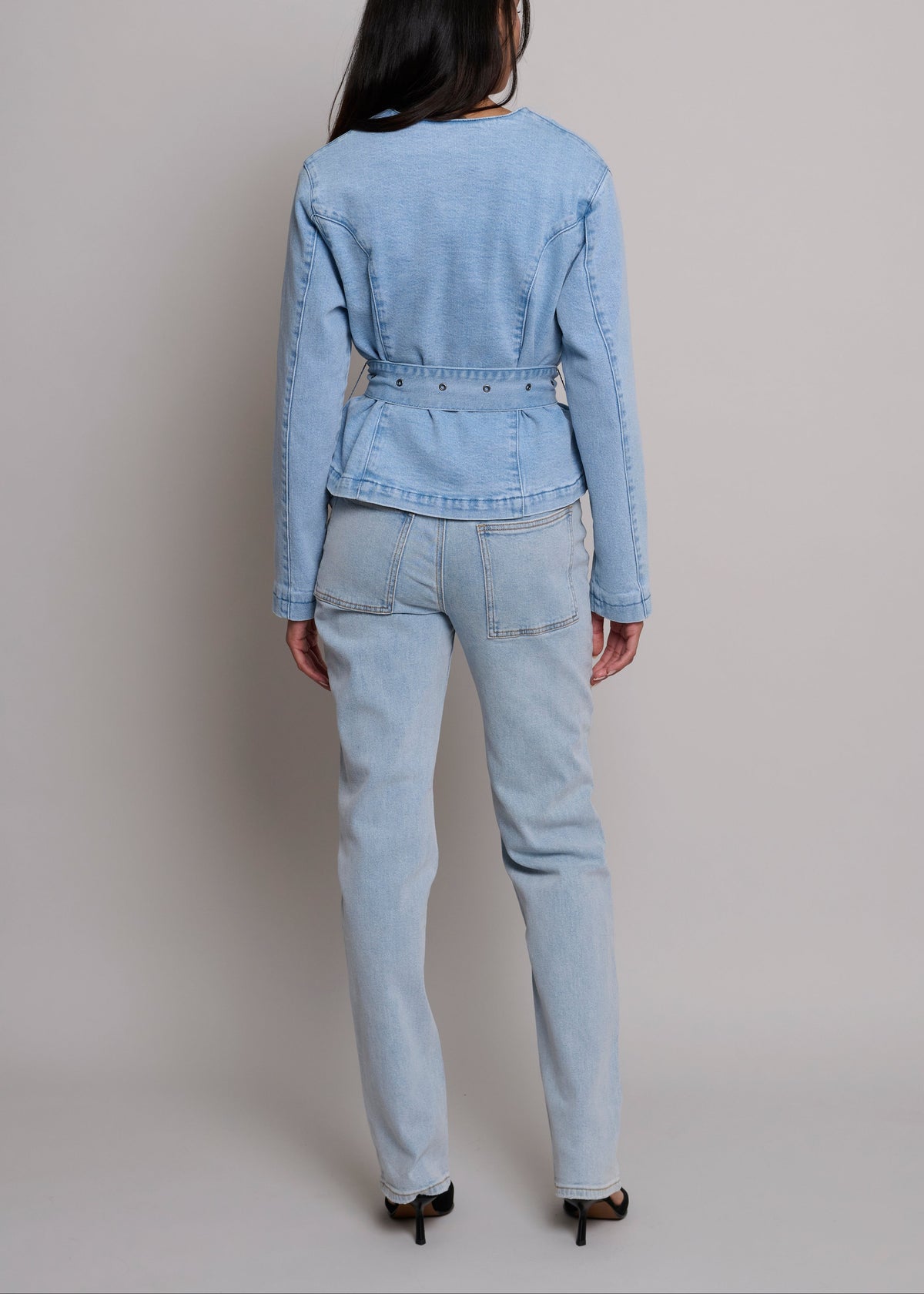 A model shown from the back wearing a light blue denim jacket and matching light blue denim jeans, paired with black high heels, against a neutral light gray studio background.