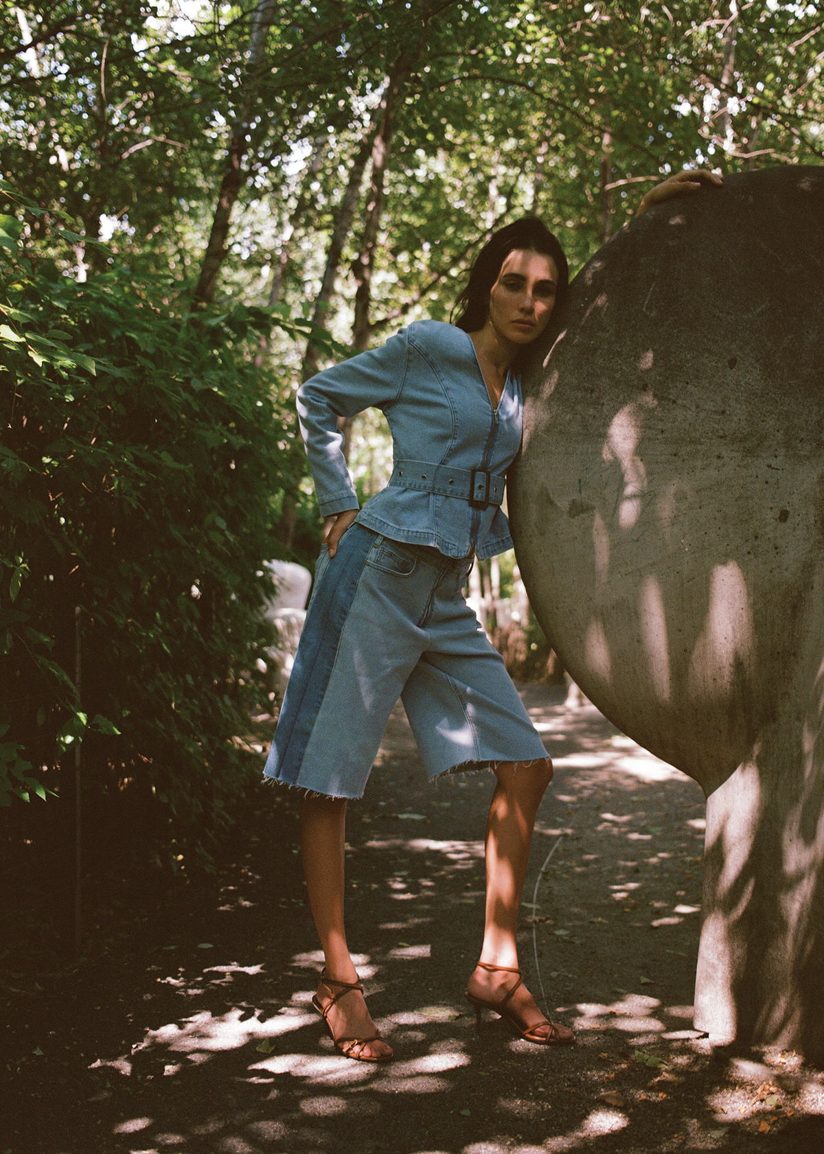 Woman in a denim outfit standing next to a large stone sculpture in a forest setting