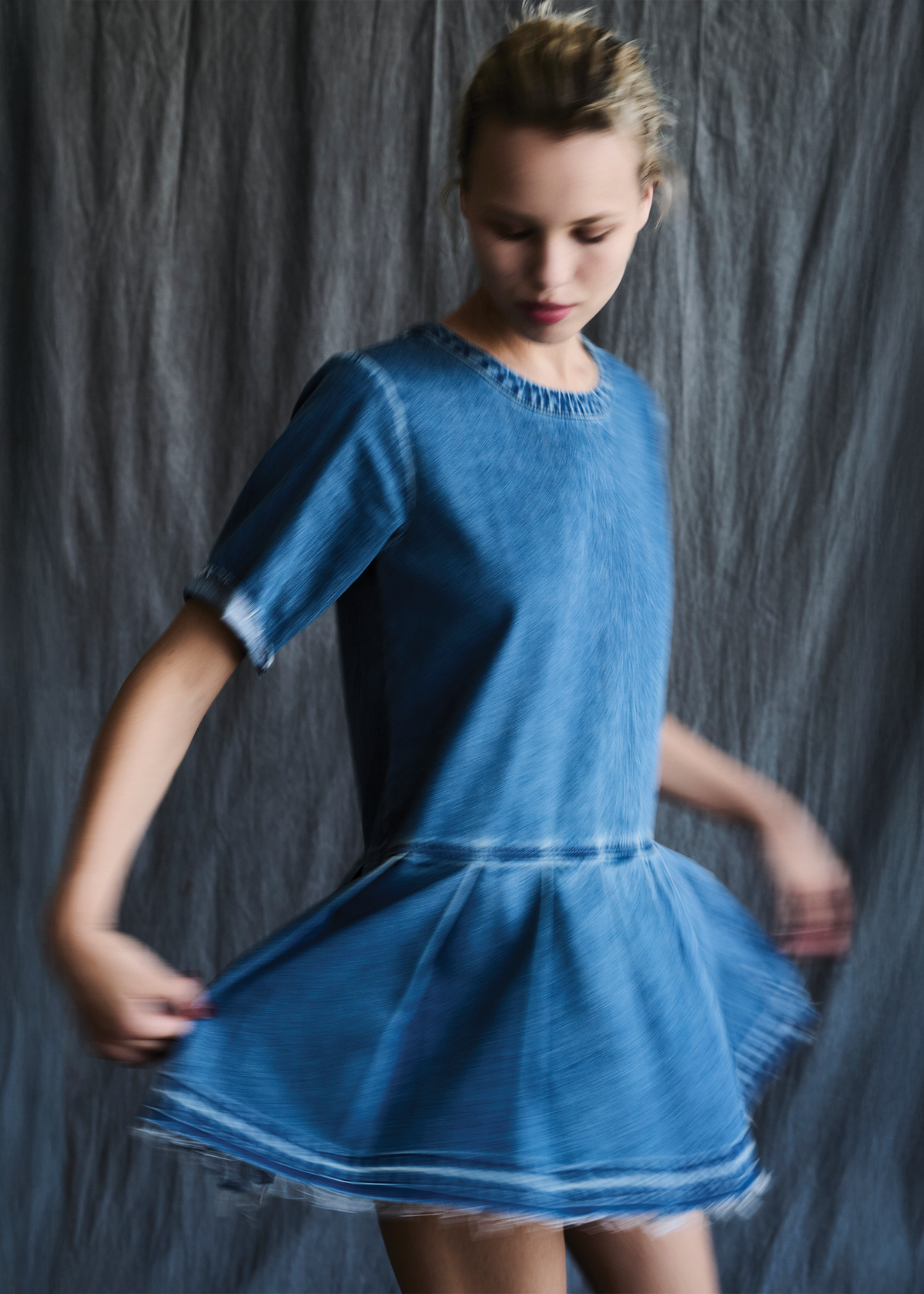 a model spinning wearing a short-sleeve denim dress with a round neckline, frayed edges, and a pleated skirt, shown against a plain white background.
