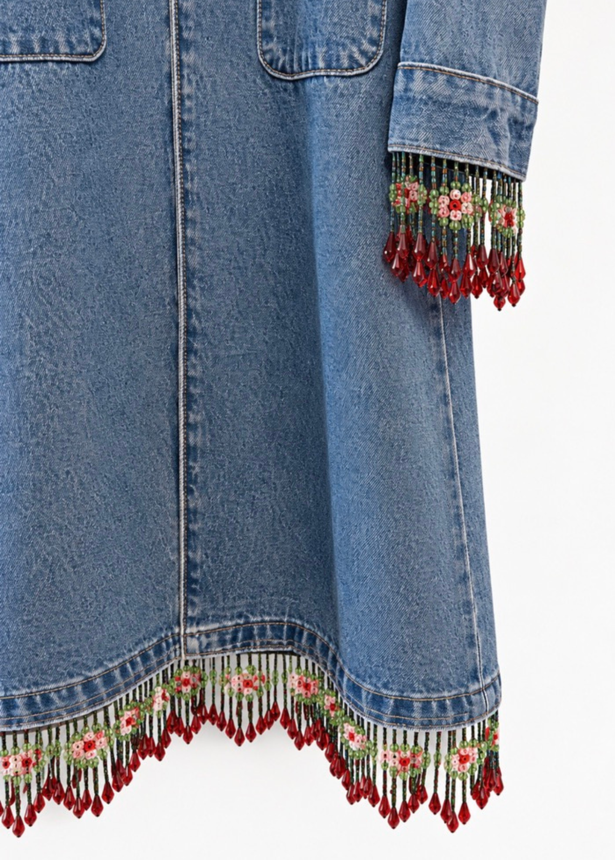 Close-up of a blue denim dress featuring floral embroidery and red beaded fringe along the hem and sleeve cuffs, shown against a plain white background.