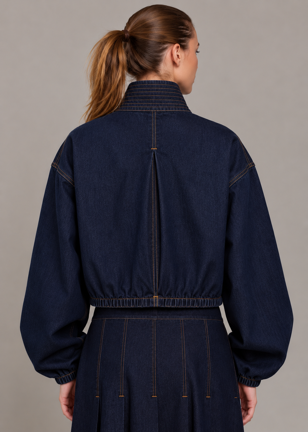 A model wearing a dark indigo denim jacket with contrast orange-brown stitching, zippered chest pockets, and a high ribbed collar, paired with a matching dark denim skirt against a neutral gray studio background.