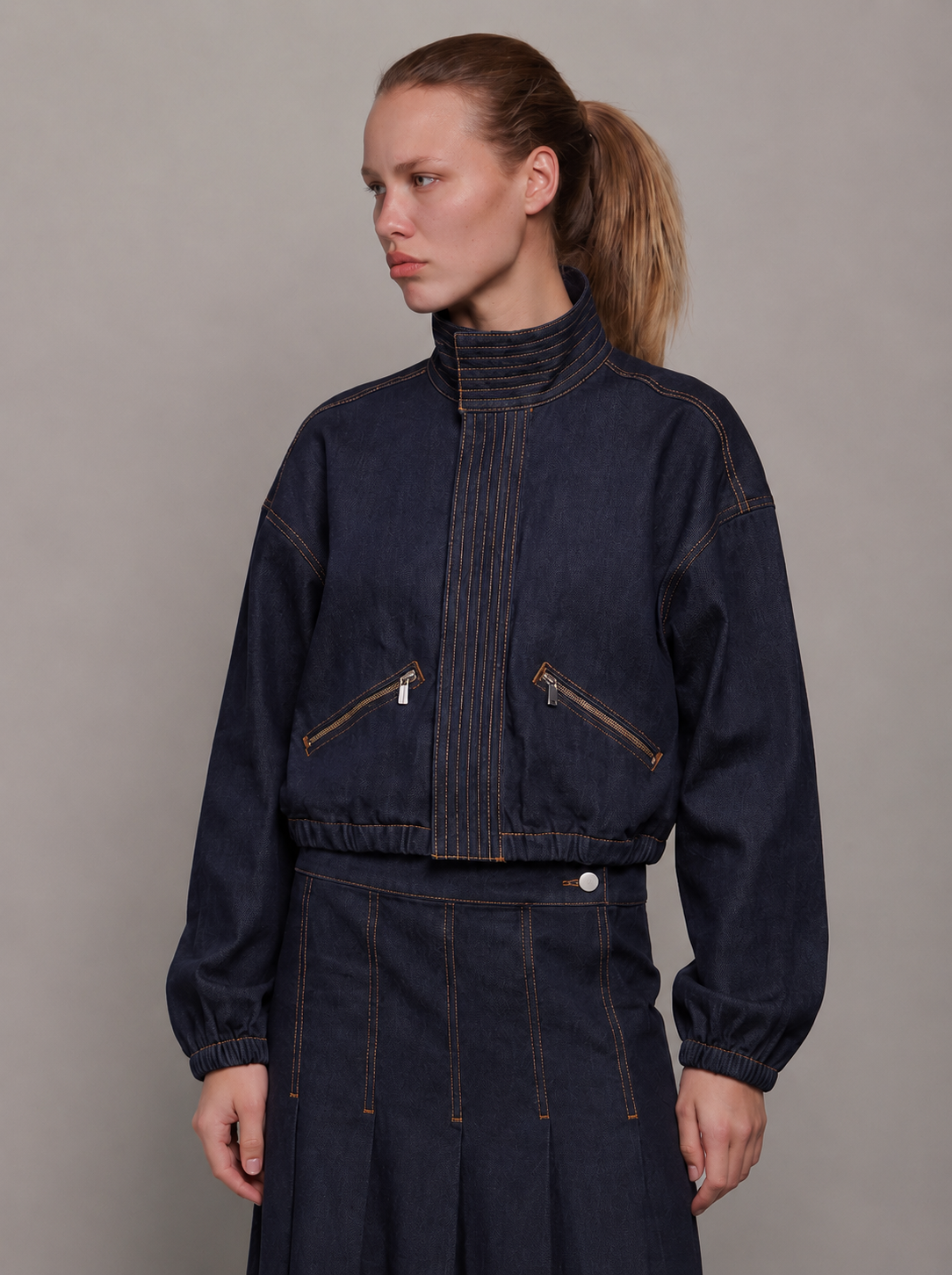A model wearing a dark indigo denim jacket with contrast orange-brown stitching, zippered chest pockets, and a high ribbed collar, paired with a matching dark denim skirt against a neutral gray studio background.